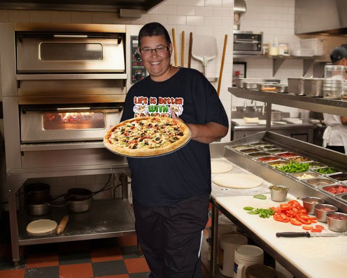 Person holding a pizza in a kitchen with various toppings and an oven in the background.